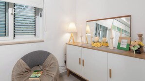 Ein Zimmer mit Fenster, Jalousien, einem Nachttisch mit Lampe, Kerzen und einem Bilderrahmen.