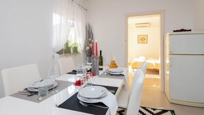 Ein Essbereich mit einem weißen Tisch, gedeckt für vier Personen, inklusive Tellern, Gläsern und Besteck. Rechts daneben befindet sich ein weißer Kühlschrank und ein Durchgang zu einem Schlafzimmer.