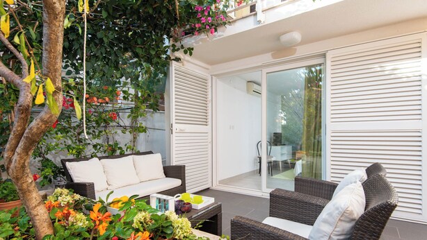 Ein Patio mit Korbmöbeln, einer weißen Couch und einem Esstisch. Eine Glastür führt in einen Innenbereich.