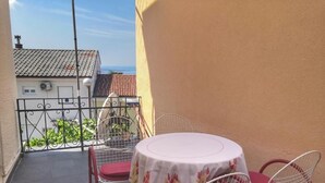 Een balkon met een ronde tafel en twee stoelen, uitzicht op een gebouw met een pannendak, en een heldere lucht.