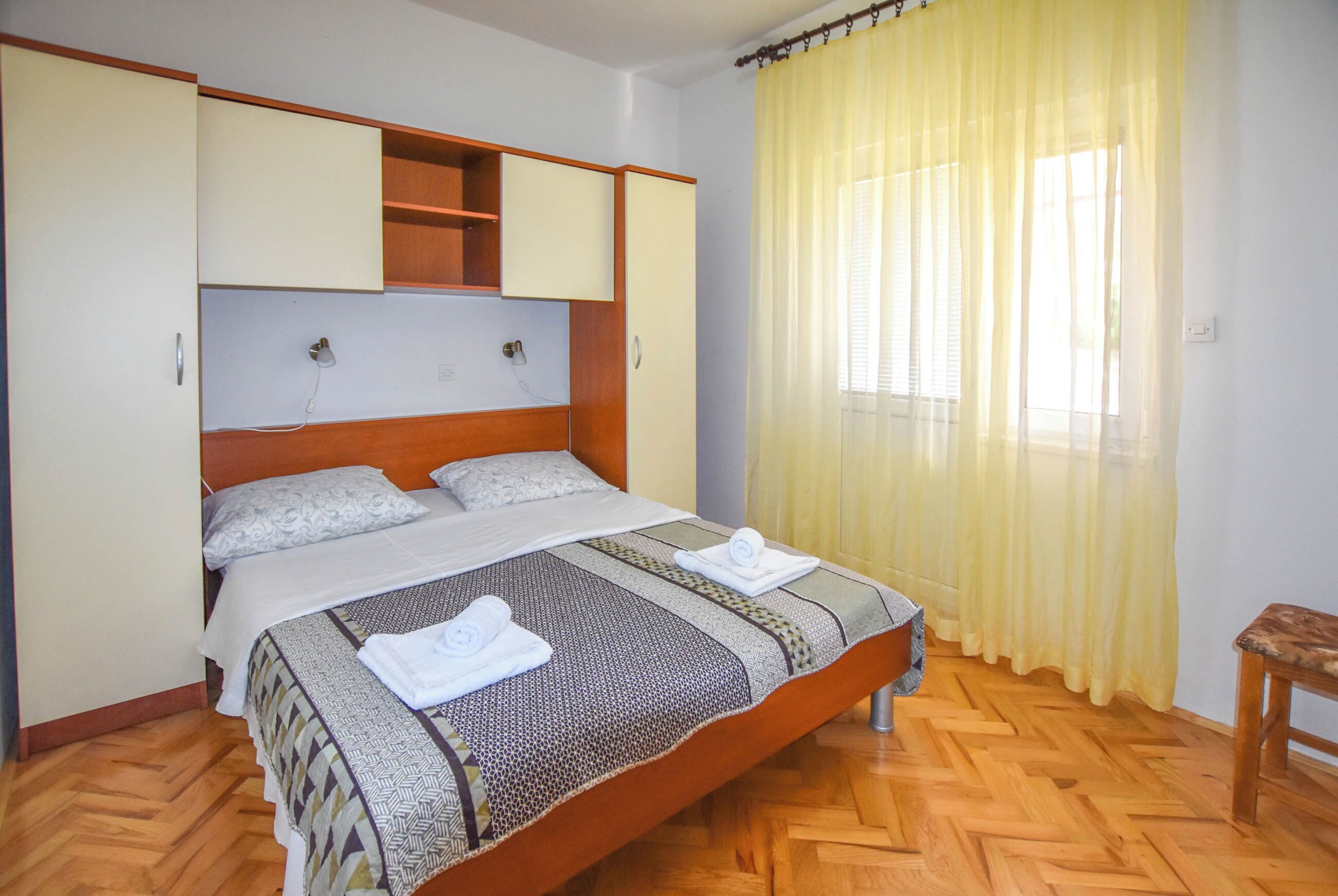 Een slaapkamer met een bed, een houten hoofdbord, een inbouwgarderobe en een raam met gele gordijnen.