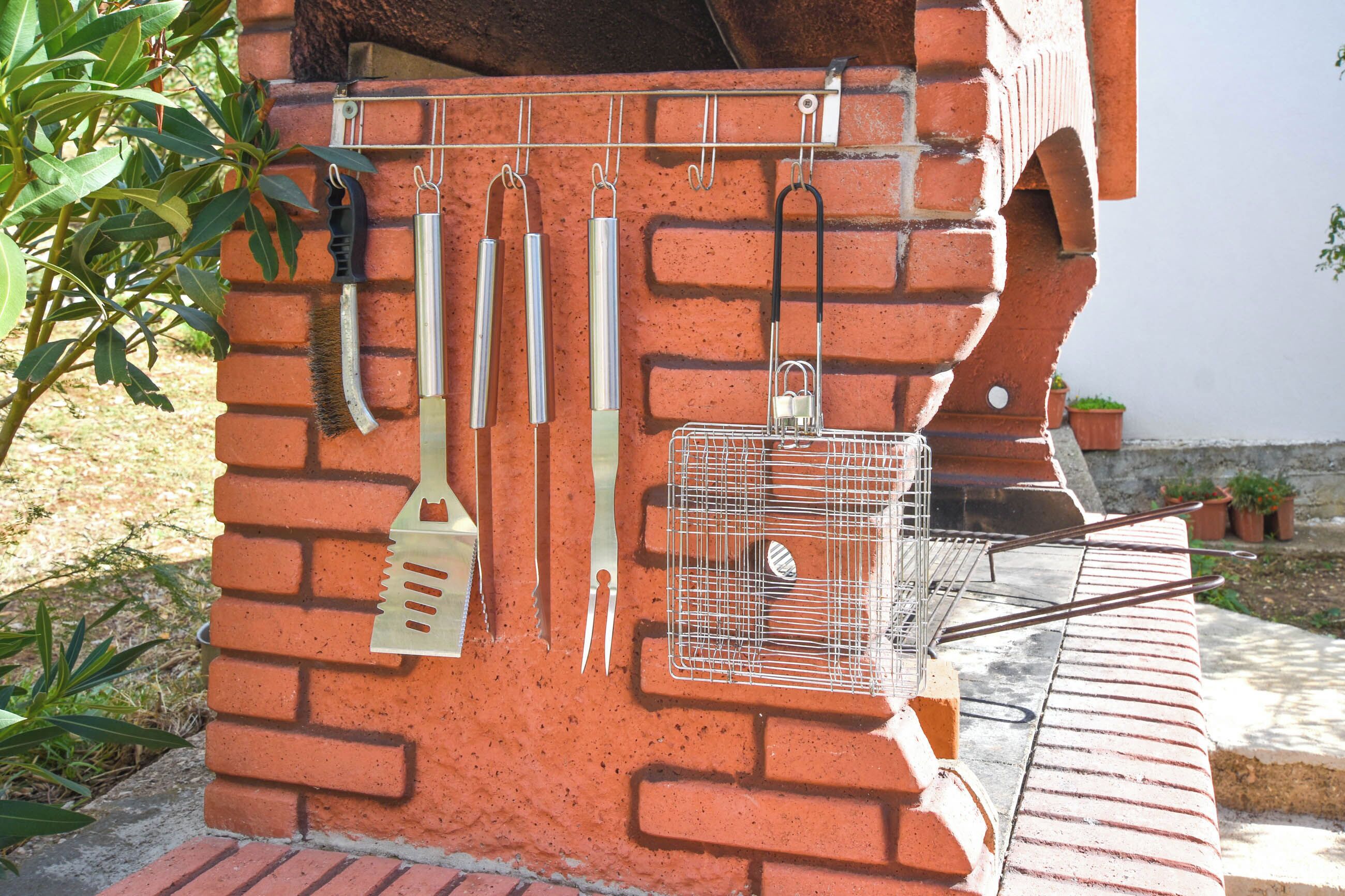 A brick barbecue grill with utensils hanging above and a wire rack below.