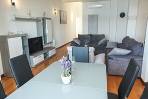 A modern living room with a grey sofa set, a dining table with a potted plant, and a TV on a stand. - Apartments Seafront (Pasman)