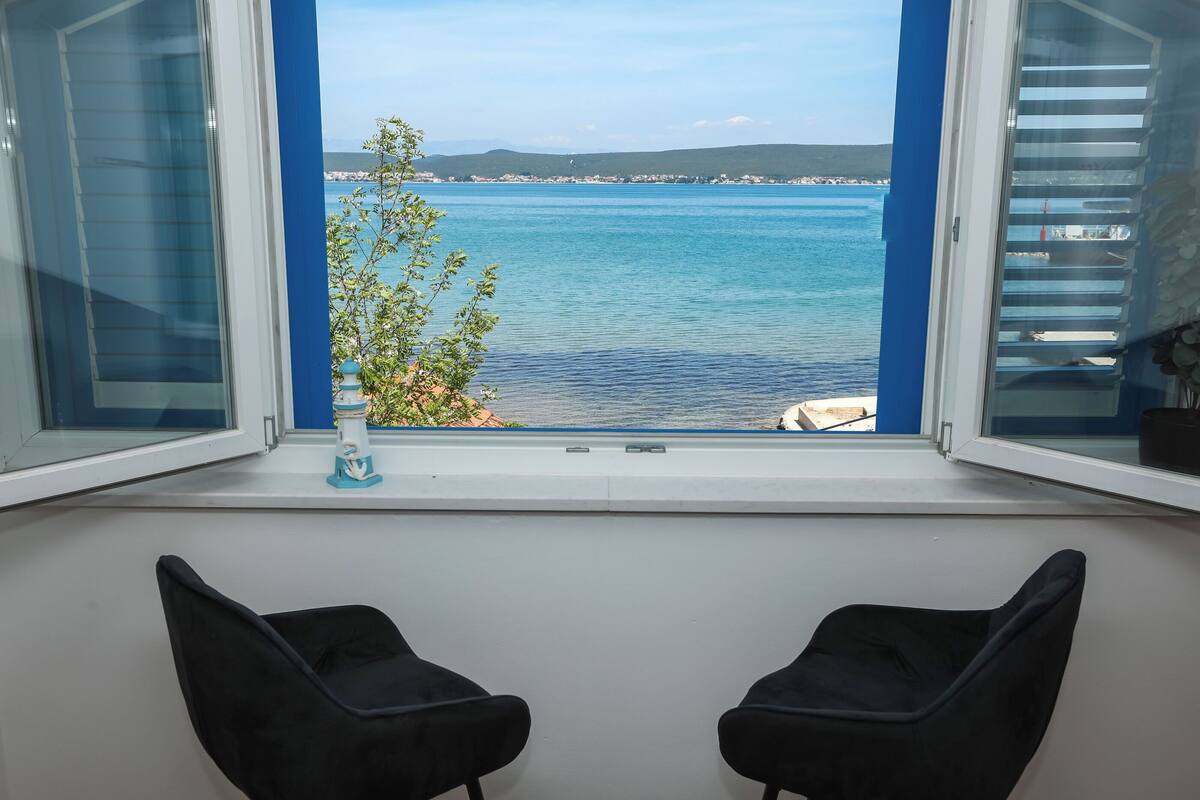Une pièce avec vue sur une mer calme et des collines lointaines. On peut voir deux chaises noires au premier plan.
