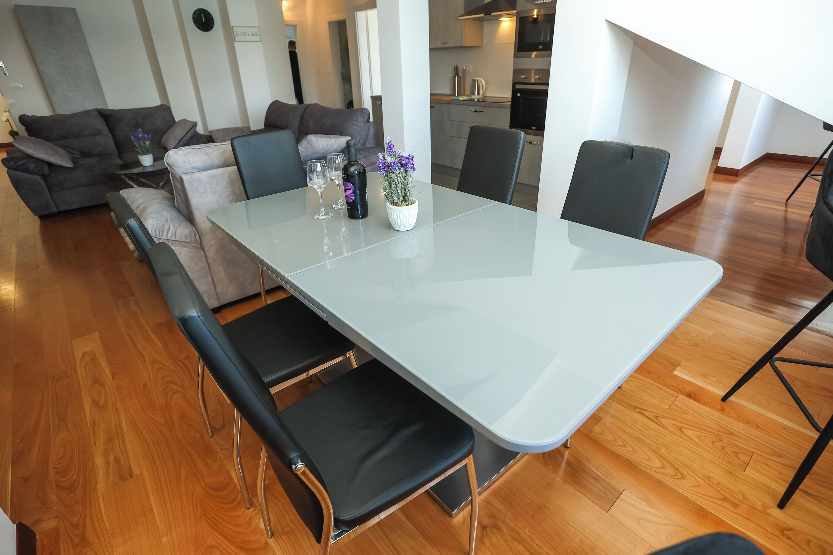 A modern living room with a grey sofa, a dining table with chairs, and a kitchen in the background.