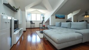 A modern loft apartment with a large gray sectional sofa, a dining area with two chairs, a kitchen with white cabinets, and a large window with blinds. - Apartments Seafront (Pasman)