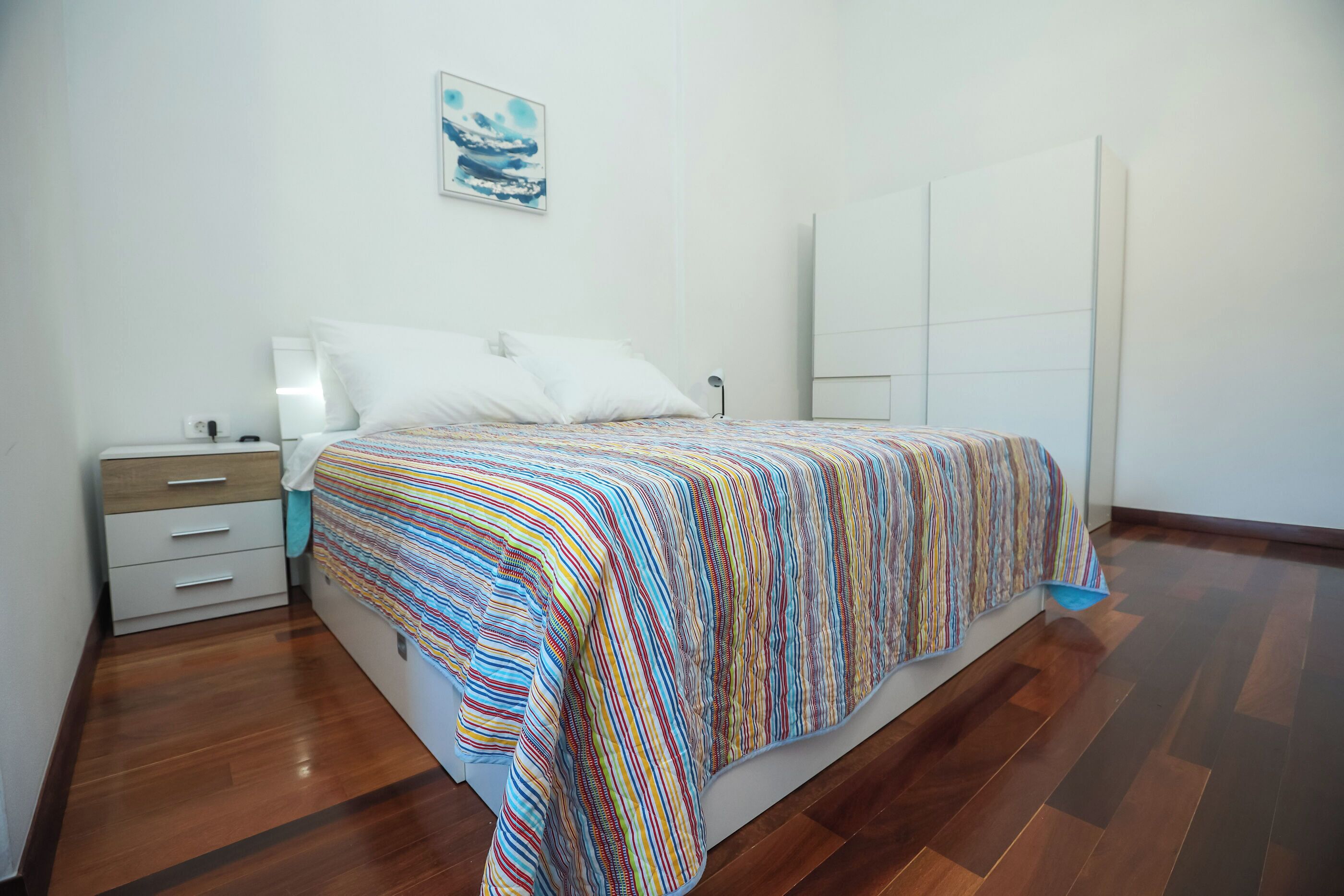 A bedroom with a bed, a nightstand, a drawer unit, a wall-mounted lamp, and a wardrobe.