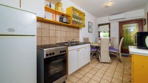 Eine kompakte Küche mit Herd, Backofen und Spüle. Gelbe Regale befinden sich über der Arbeitsfläche, und es gibt einen Essbereich mit Tisch und Stühlen.