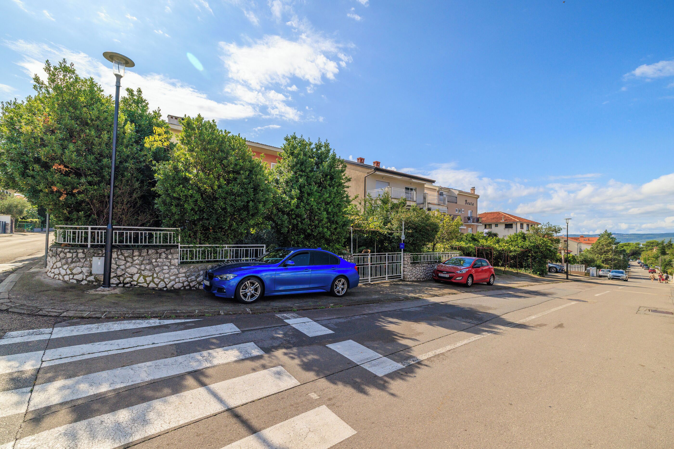 A street view with a blue car parked by the curb, a red car in the background, and a pedestrian crossing.