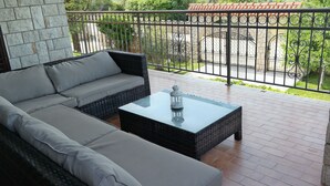 Un patio al aire libre con un sofá, una mesa de vidrio, un farol y un muro de piedra al fondo.