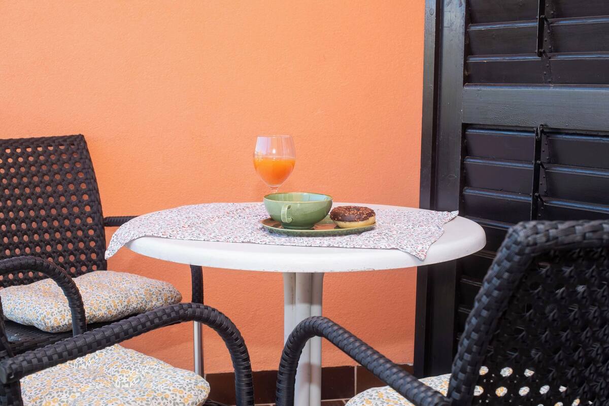 Una mesa redonda pequeña con un bol verde y un vaso de zumo de naranja, rodeada de sillas de mimbre con cojines estampados.