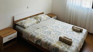 A bedroom with a bed, wooden nightstands, a bedside table, and a window with sheer curtains. - Apartments Vladimir (Malinska-Dubasnica)