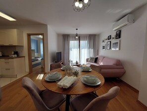 A modern living room with a dining table set for four, a pink sofa, and a kitchen area visible through an open door. - Apartment Crikva (Crikvenica)