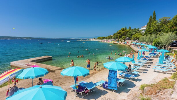Une plage avec une eau d’un bleu limpide, des gens qui se baignent et des transats avec des parasols.