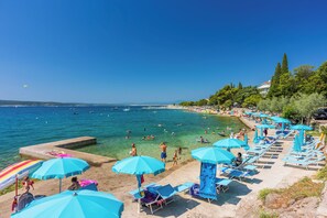 A beach with clear blue water, people swimming, and sun loungers with umbrellas. - Apartment Crikva (Crikvenica)