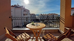 Un balcon avec une table ronde en bois et deux chaises en bois, donnant sur un quartier résidentiel avec des maisons et un ciel dégagé.