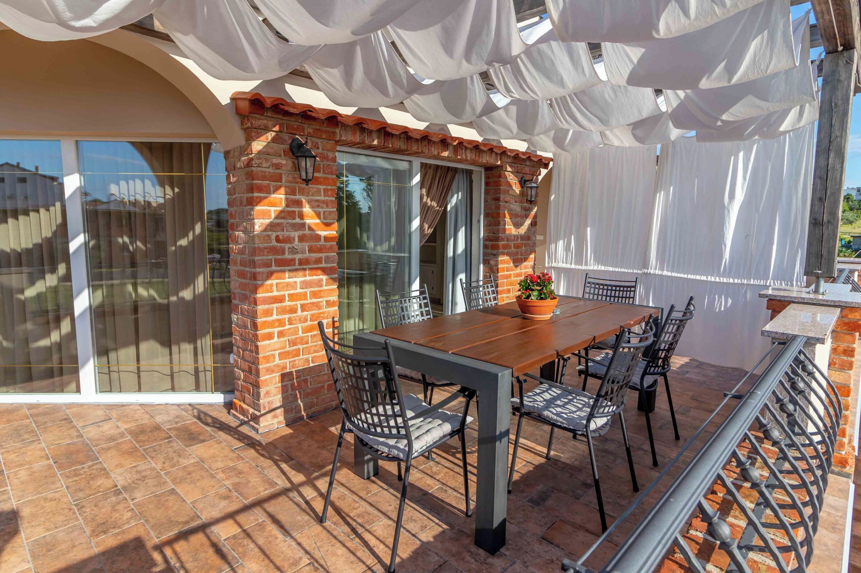 Eine Terrasse im Freien mit einem Holztisch und Stühlen, einer Ziegelwand und einer Pergola mit weißem Stoff.