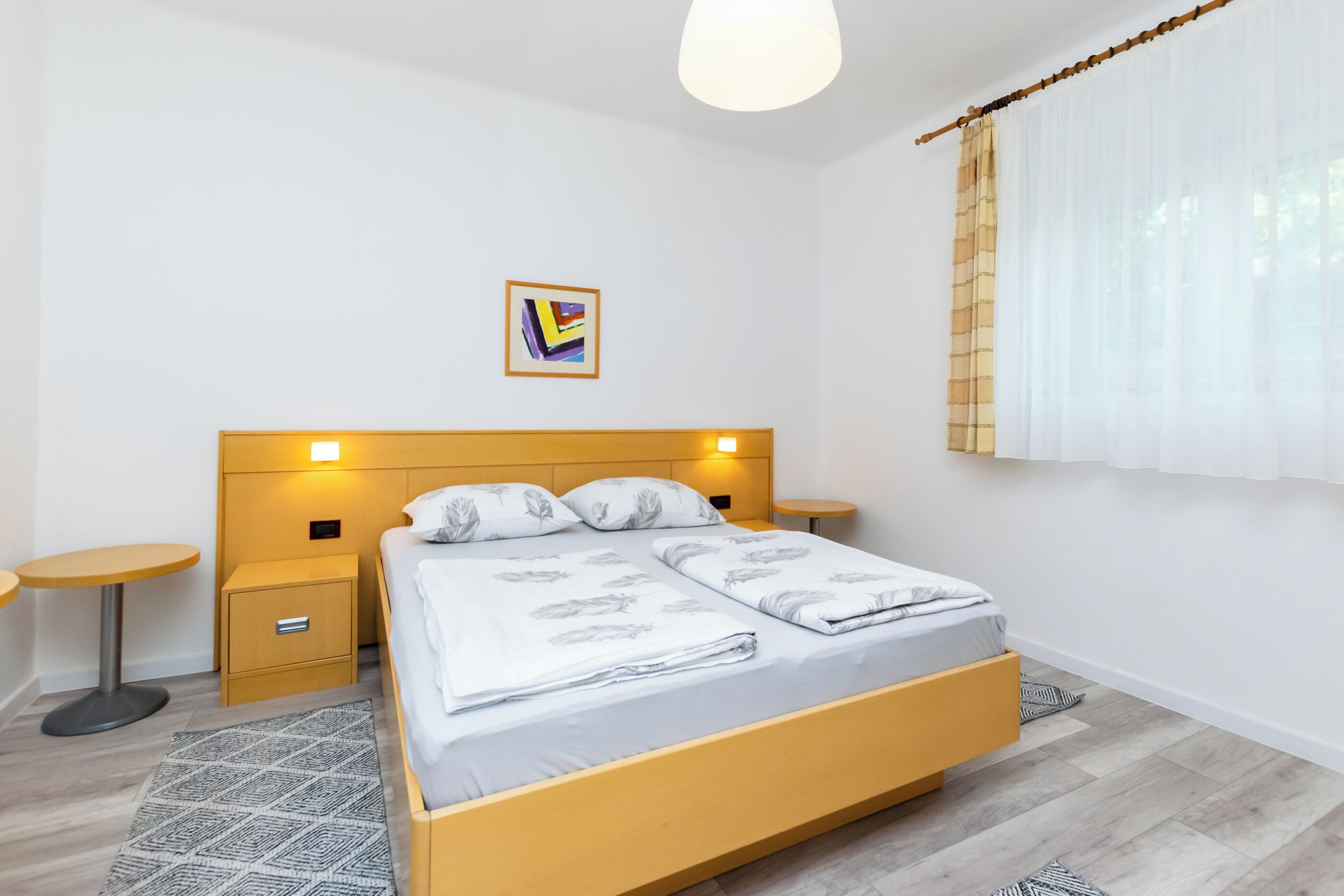 A modern bedroom with a wooden bed, bedside tables, a small round table, and a wall-mounted light fixture.