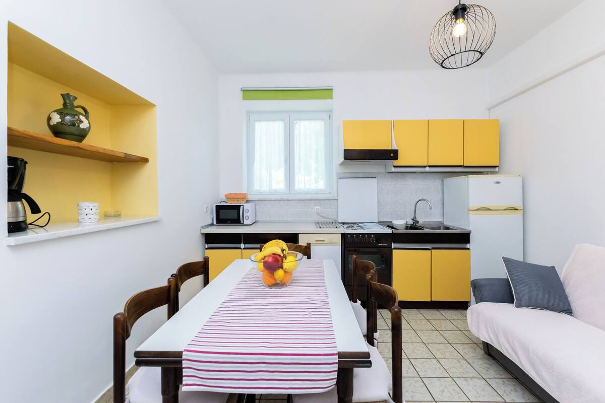 A compact kitchen with yellow cabinets, a white countertop, and a dining area with a striped tablecloth.