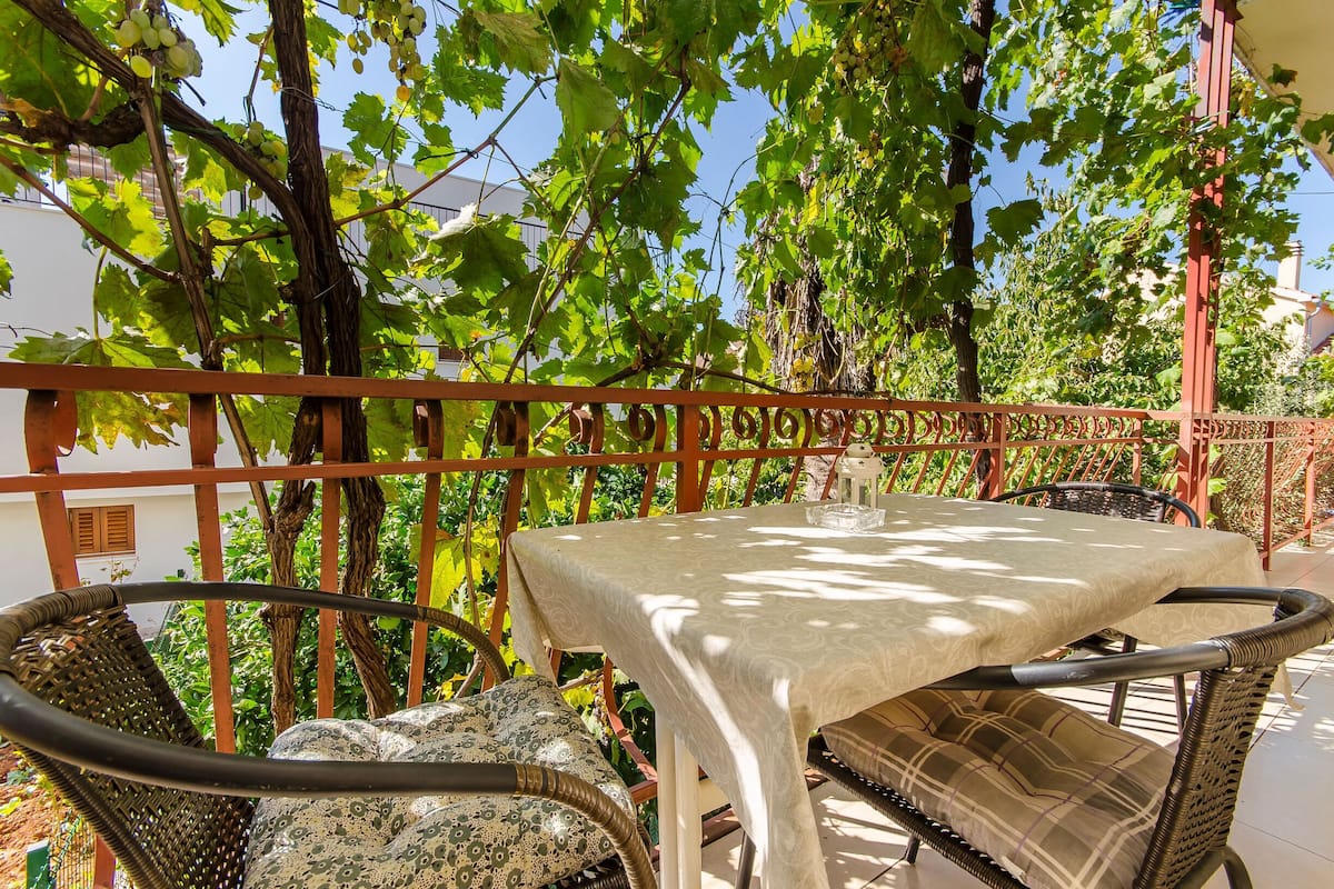 Eine Terrasse mit Tisch und Korbstühlen, umgeben von Grünpflanzen und einem roten Geländer.