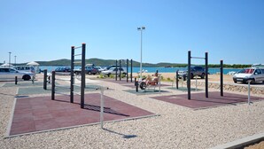 Un espace de remise en forme en plein air, équipé d’appareils d’exercice, un parking et une vue panoramique sur la mer et les collines en arrière-plan.