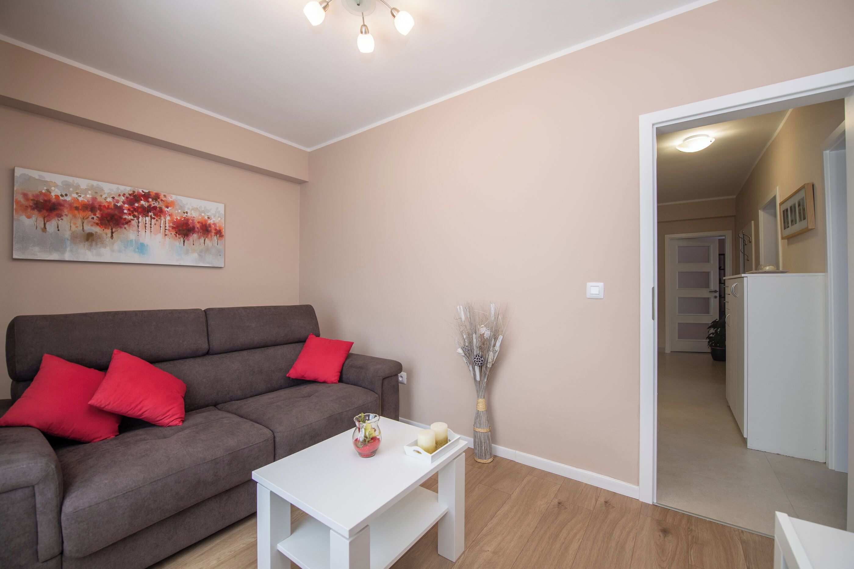 Ein Wohnzimmer mit grauem Sofa, roten Kissen, einem weißen Couchtisch, einer Vase mit Trockenpflanzen und einem Gemälde an der Wand.