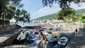 Eine Küstenansicht mit einer Statue einer sitzenden Person, Booten, die an der Pier festgemacht sind, und einer Stadtansicht im Hintergrund.