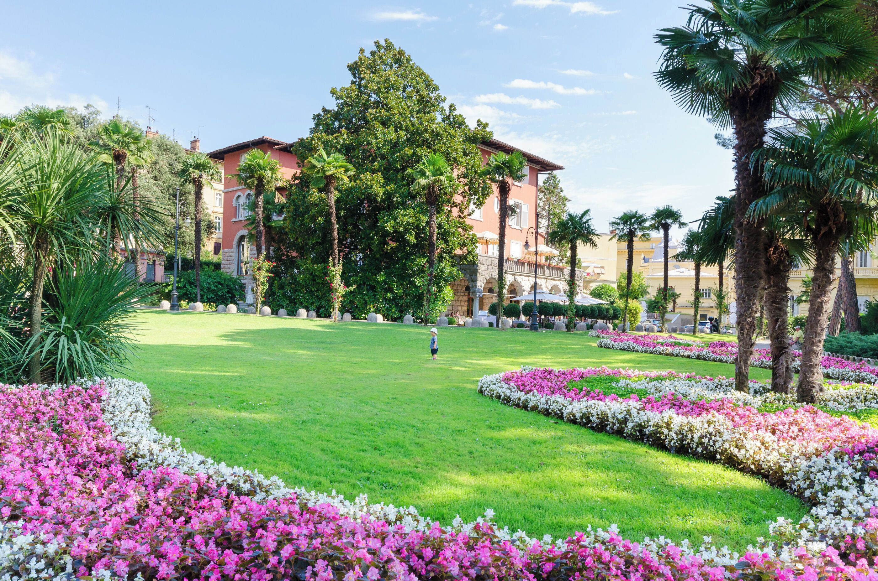 Un jardin bien entretenu, avec une pelouse centrale, bordé de parterres de fleurs et de palmiers, qui mène à un bâtiment à la façade rouge et aux fenêtres en arc de cercle.