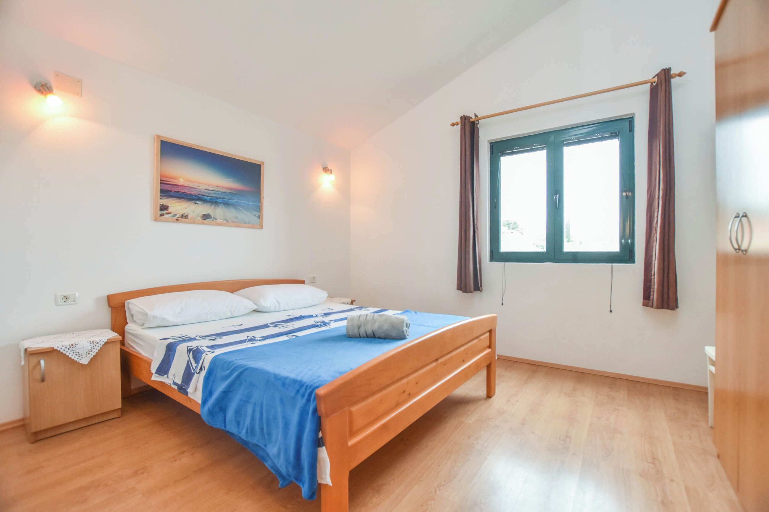 A bedroom with a wooden bed, a nightstand, a painting on the wall, a window with curtains, and a light fixture.
