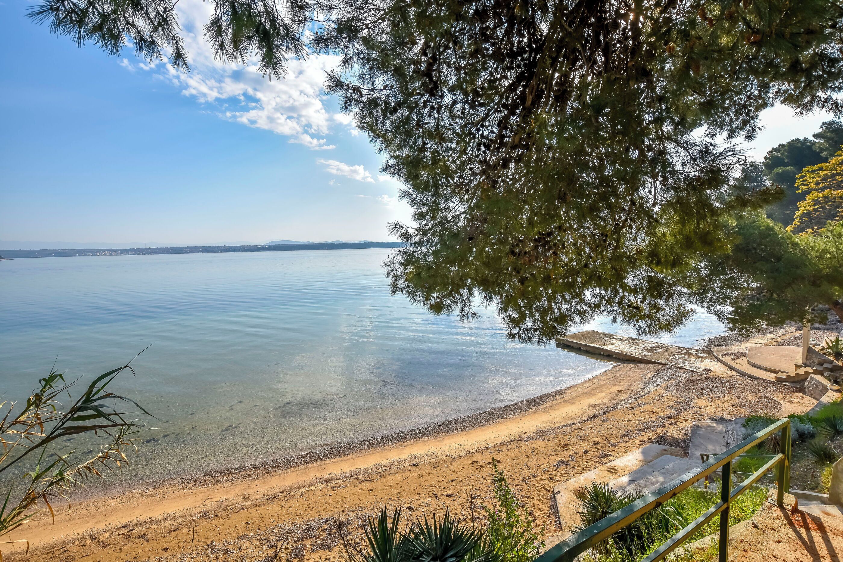 Une plage à l’eau claire, avec un rivage sablonneux et un sentier pavé de pierres menant à un bâtiment.