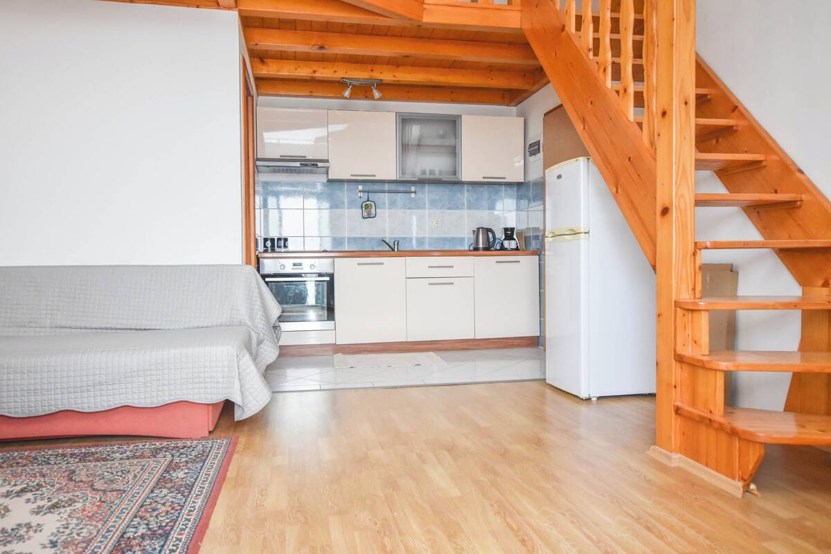 Een kleine keuken met witte kastjes, een houten trap en een bed met een beddegoed met een patroon.