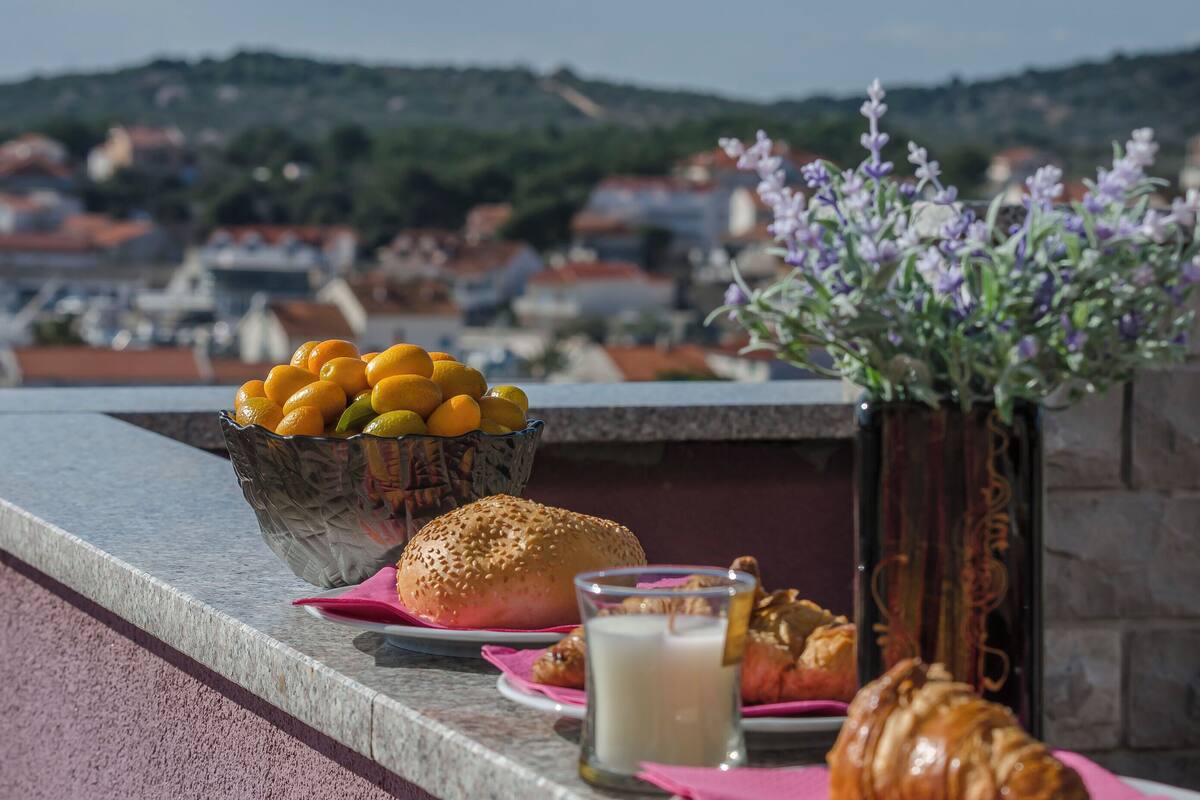 A table with a bowl of lemons, a sandwich, a glass of milk, and a croissant on a balcony overlooking a town.