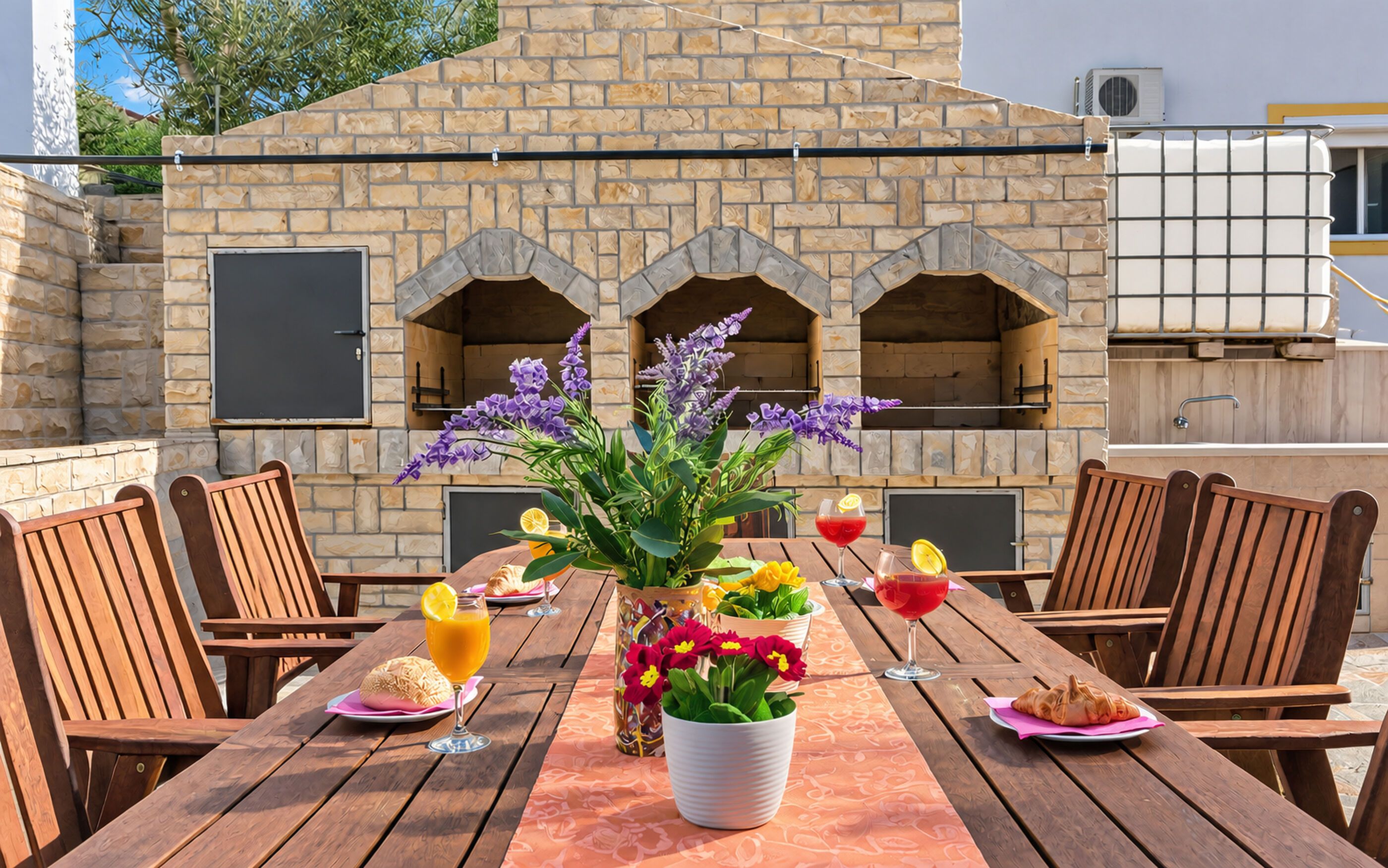 An outdoor dining area with a stone wall and arched openings, wooden chairs, a table with a vase of flowers, and plates with food and drinks.