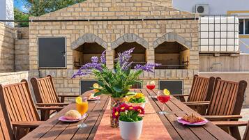 An outdoor dining area with a stone wall and arched openings, wooden chairs, a table with a vase of flowers, and plates with food and drinks.