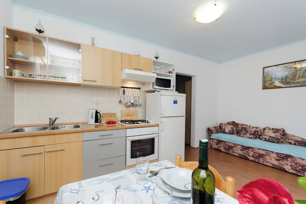 A compact kitchen with a microwave, oven, and sink, a dining table set with plates and a bottle, and a sofa in the adjacent living area.