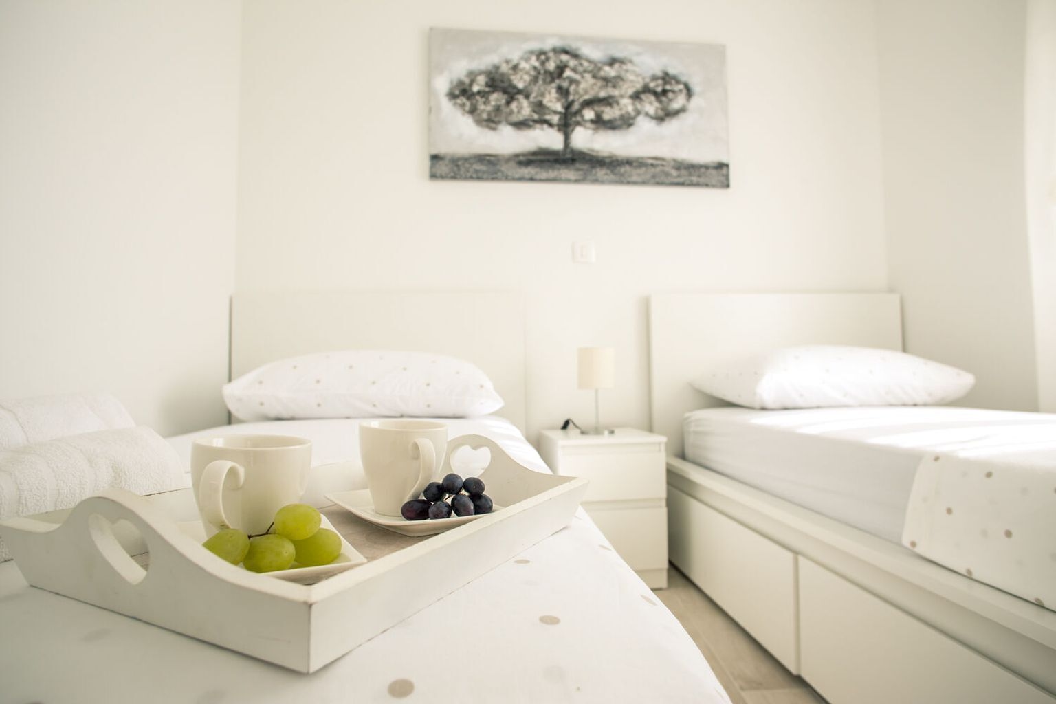 Una habitación de hotel con una cama, una mesita de noche con una lámpara, una bandeja con frutas y tazas, y un cuadro de un árbol en la pared.