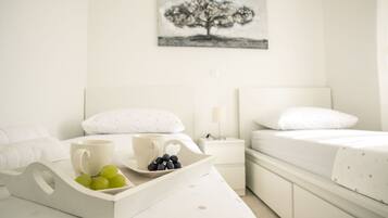 Una habitación de hotel con una cama, una mesita de noche con una lámpara, una bandeja con frutas y tazas, y un cuadro de un árbol en la pared.