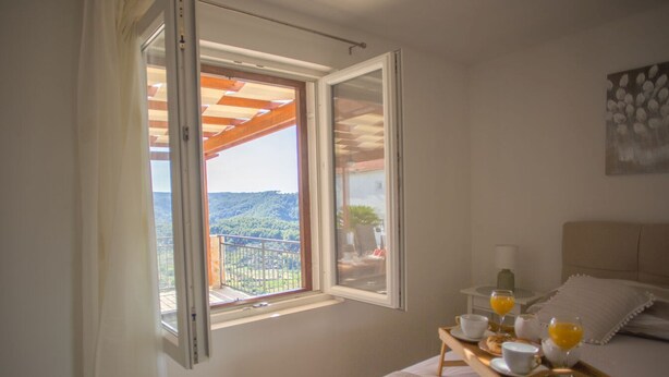 Une chambre avec un lit, une table de chevet sur laquelle se trouve un plateau avec des articles pour le petit-déjeuner, une fenêtre avec un volet ouvert, et une vue sur un balcon et la verdure environnante.