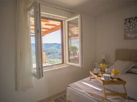 Un dormitorio con cama, una mesita de noche con una bandeja de desayuno, una ventana con persiana abierta y vistas a un balcón y follaje exterior.
