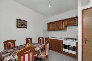 A kitchen with wooden cabinets, a gas stove, and a tiled backsplash. There is a dining table with chairs and a fruit bowl. - Apartments Robinzon Nikola (Sucuraj)