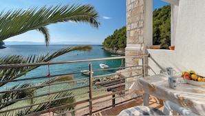 Ein Balkon mit Blick auf das Meer, ein Tisch mit Obstschale und ein Fenster mit Ausblick auf eine bewaldete Gegend.