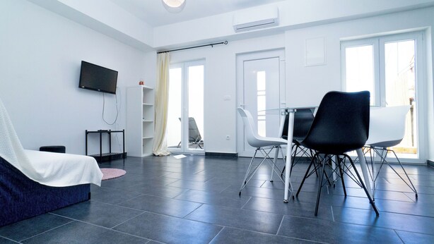 A modern hotel room with a bed, a TV mounted on the wall, a dining area with chairs, and a large window. - Apartments Annela (Seget)