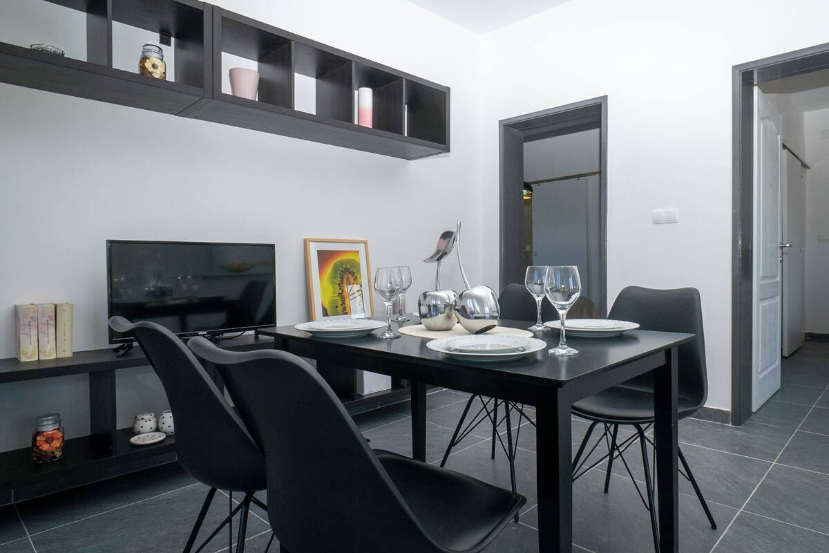 A modern dining area with a dark table set for two, black chairs, a television, and a shelf with decorative items.