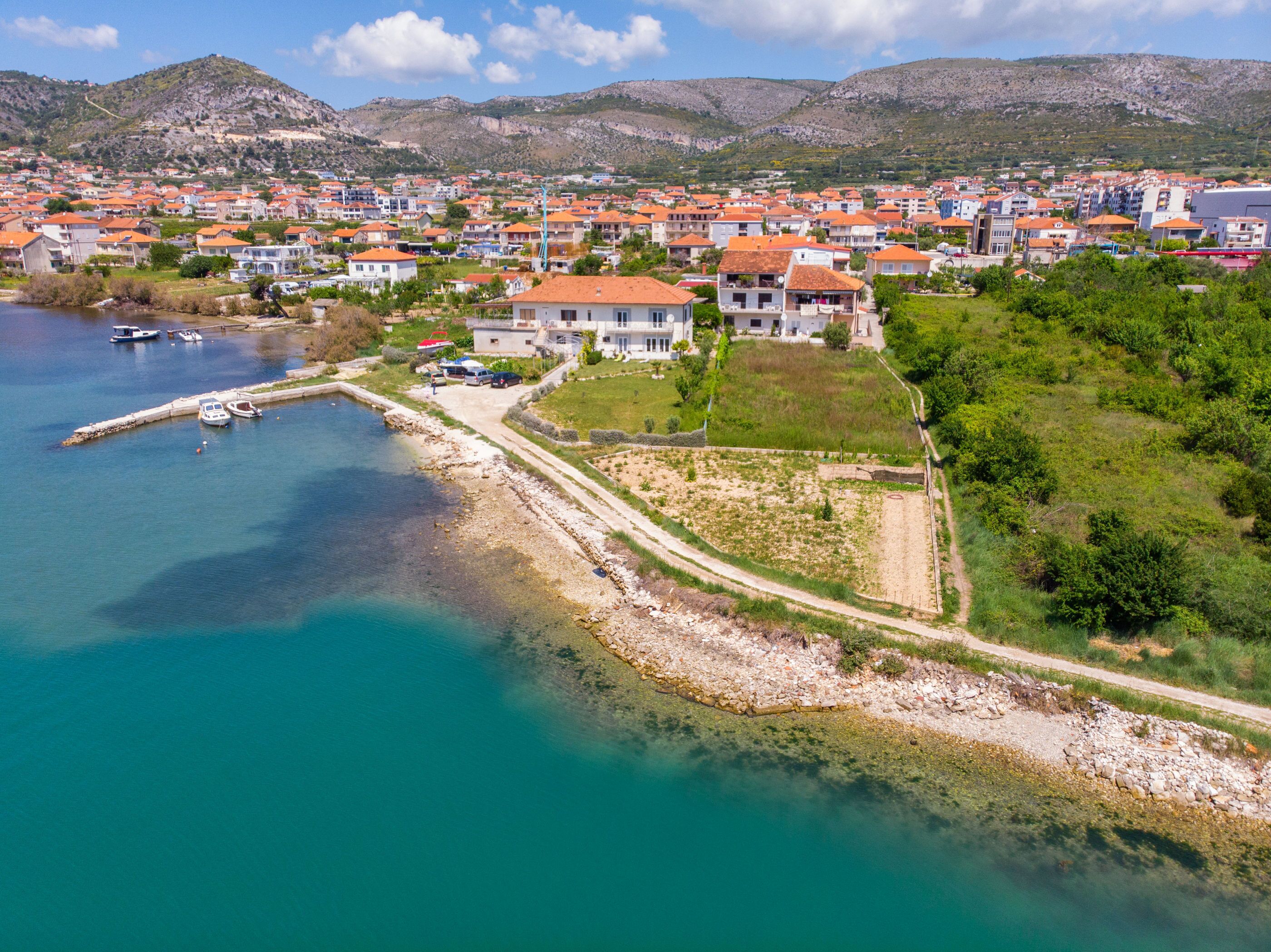 An aerial view of a coastal town with houses, a marina, and a road leading to a large house with a garden.