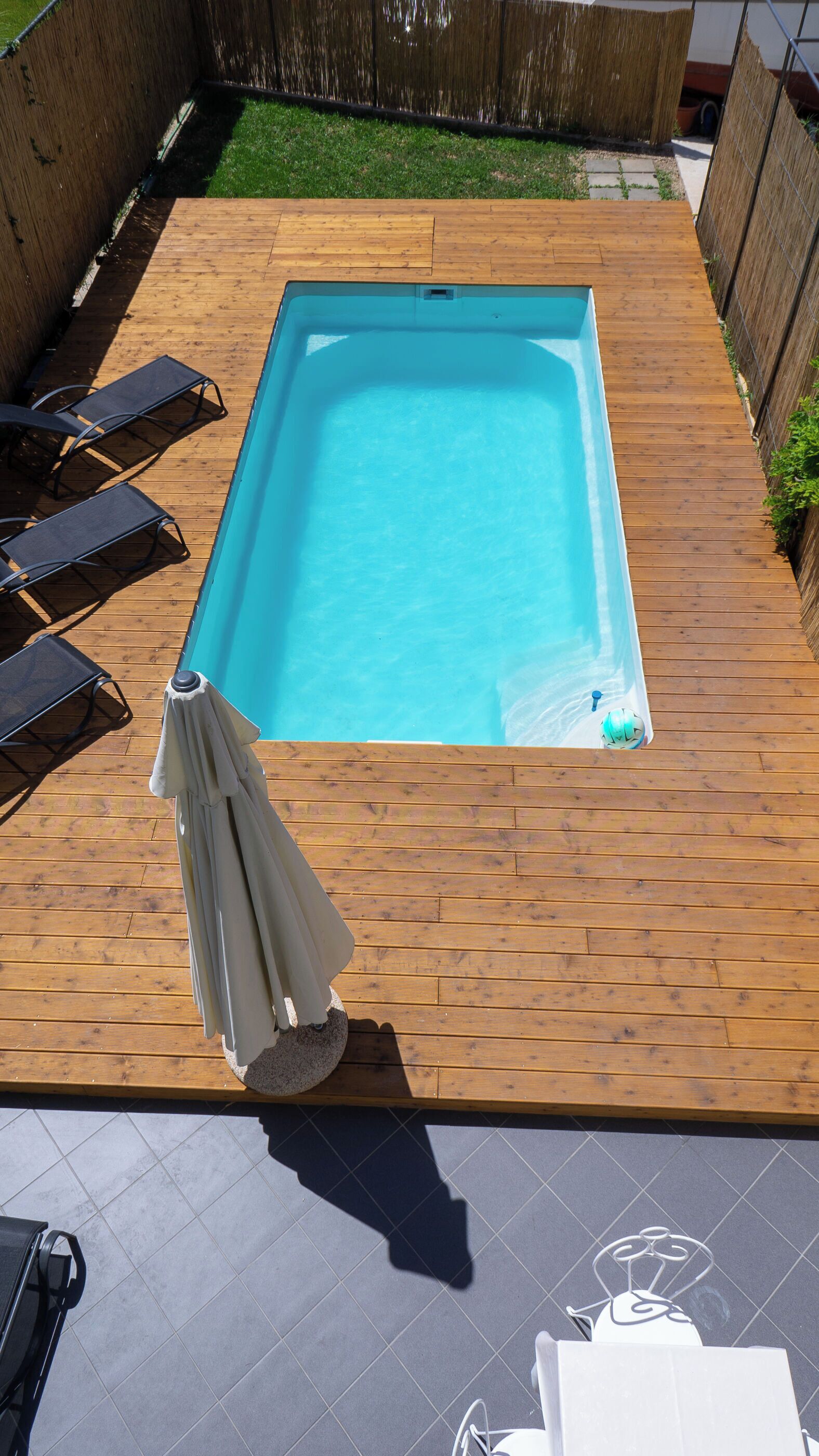 Une piscine rectangulaire entourée d’une terrasse en bois, avec un parasol blanc et plusieurs chaises longues.