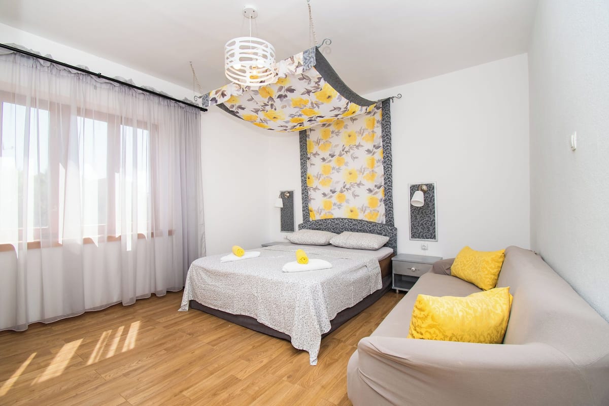 A bedroom with a bed, a bedside table, a lamp, a sofa, and a window with curtains.