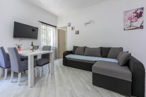 A modern living room with a white dining table, grey chairs, a black sofa, a flat-screen TV, and a window with sheer curtains. - Apartment Danijel (Novi Vinodolski)