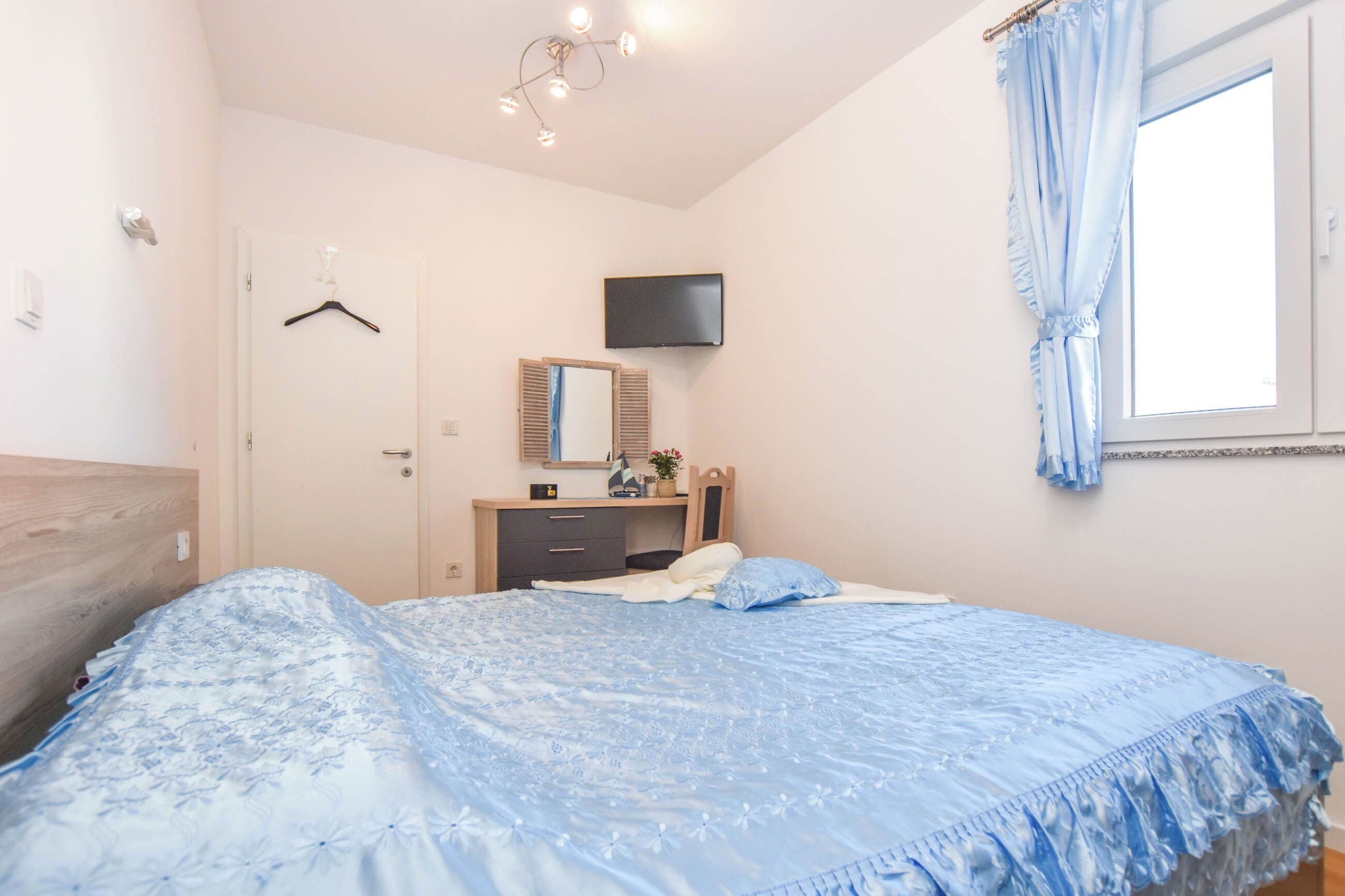 Un dormitorio con cama, cómoda, espejo, televisor y ventana con cortinas.