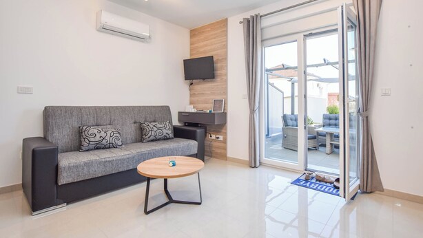Un soggiorno moderno con un divano grigio, un tavolino in legno, una televisione a schermo piatto montata su un pannello di legno e una porta scorrevole in vetro che dà su un'area relax all'aperto.