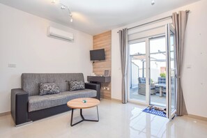 A modern living room with a grey sofa, a wooden coffee table, a flat-screen TV mounted on a wooden panel, and a sliding glass door leading to an outdoor seating area. - Apartment Martin (Biograd na Moru)